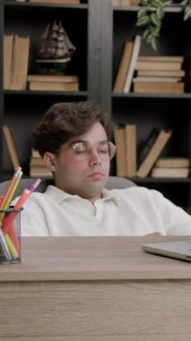 Gözlüklü bir erkek öğrenci masanın arkasına yaslanıyor, dizüstü bilgisayara bakıyor, arka planda kitaplıklar ve bitkiler var.