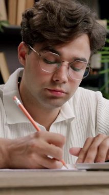 Kıvırcık saçlı genç adam ahşap masadaki kalemle notlar yazmaya odaklanıyor. Etrafı kırtasiye malzemeleri ve rahat çalışma alanındaki bitkilerle çevrili.