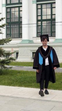 Mezuniyet kıyafetli bir erkek öğrenci kaldırımda yürüyor, neşeyle mezuniyet şapkasını kaldırıyor üniversitenin dışında yeşil ağaçlar ve klasik mimariyle.