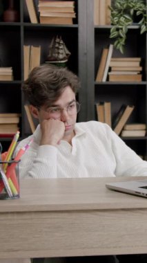 Gözlüklü bir erkek öğrenci bir masada otururken, bir dizüstü bilgisayara odaklanıyor, kitaplıklar ve kırtasiye ile çağdaş bir çalışma ortamında görülebilir