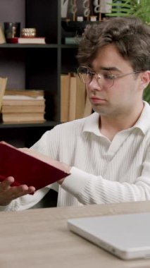 Kıvırcık saçlı bir erkek öğrenci, ahşap bir masada, kitap rafları ve sıcak bir çalışma ortamı yaratan yeşil bir bitkiyle bir kitabı inceliyor.