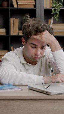 Kahverengi saçlı erkek öğrenci, önce hayal kırıklığı, sonra yorgunluk ve son olarak rahat bir kütüphane ortamında masa başında çalışırken rahatlama sergiliyor.