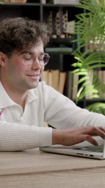 Kıvırcık saçlı öğrenci, rahat bir çalışma ortamında, kitap rafları ve yeşil bitkilerle çevrili, ahşap bir masadaki dizüstü bilgisayarda yazmaya odaklanıyor.