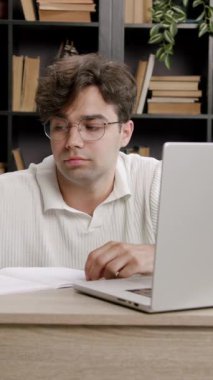 Gözlüklü bir erkek öğrenci, kitaplarla dolu samimi bir kütüphane ortamında dizüstü bilgisayar ve kağıtlarla çalışırken masasında hayal kırıklığına uğramış görünüyor.