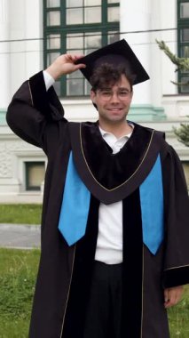 Mezuniyet kıyafetli bir erkek öğrenci üniversitenin dışında ağaçlar ve arka planda görünen bir binayla şapkasını havaya fırlatıyor.