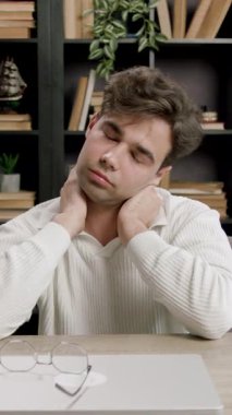 Beyaz kazaklı erkek öğrenci, kitaplıklar ve çalışma materyalleriyle çevrili bir masada boyun ve sırt germe hareketleri yapıyor.