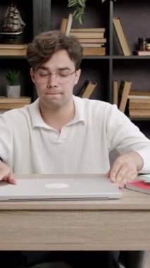 Beyaz kazaklı genç bir adam çalışma materyallerini bir masanın üzerine yerleştiriyor. Evrakları ve kırtasiyeleri, arkasında kitaplıklar olan parlak bir ev ofisinde taşıyor.