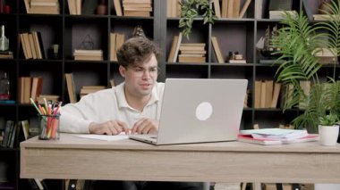 Gözlüklü öğrenci, kitaplarla ve kırtasiyelerle çevrili bir masada dizüstü bilgisayarla meşgul oluyor. Çalışma sırasında odak noktasından düşünceye geçiş gösteriyor.