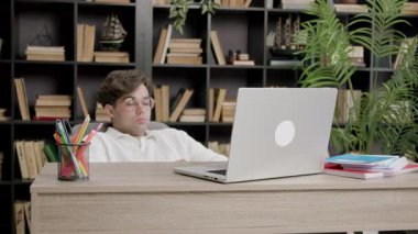 Gözlüklü bir erkek öğrenci masanın arkasına yaslanıyor, dizüstü bilgisayara bakıyor, arka planda kitaplıklar ve bitkiler var.