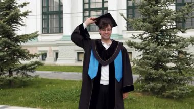 Mezuniyet kıyafetli bir erkek öğrenci üniversitenin dışında ağaçlar ve arka planda görünen bir binayla şapkasını havaya fırlatıyor.