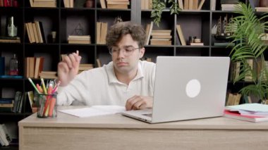 Gözlüklü bir erkek öğrenci, kitaplarla dolu samimi bir kütüphane ortamında dizüstü bilgisayar ve kağıtlarla çalışırken masasında hayal kırıklığına uğramış görünüyor.
