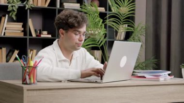 Kıvırcık saçlı ve gözlüklü genç adam ahşap bir masadaki dizüstü bilgisayarda yazmaya odaklanıyor. Etrafı kitaplarla çevrili ve çalışma odasındaki kapalı bitkilerle dolu.
