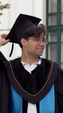 Mezuniyet kıyafetli bir erkek öğrenci açık havada seremoni için hazırlanıyor, kep ve cübbesini üniversite binası ve ağaçlarının önünde düzeltirken gülümsüyor.