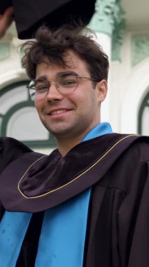 Mezuniyet kıyafetli bir erkek öğrenci, üniversitenin önünde şapkasını neşeyle sallıyor ve arkasındaki binanın etkileyici mimarisini gözler önüne seriyor.