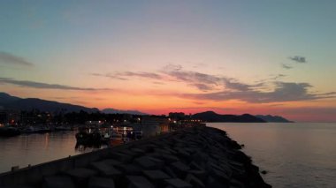 Hava perspektifi günbatımında huzurlu kıyı şeridini yakalar, sakin suları ve dağların siluetlerini aydınlatır, pürüzsüz bir kamera tavasıyla manzaranın güzelliğini arttırır.