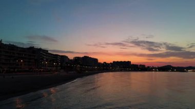 Hava perspektifi günbatımında sakin bir plajı gözler önüne seriyor. Hafif dalgalar kıyıya vuruyor. Alacakaranlık ve kentsel ışıkların güzelliğini vurguluyor.
