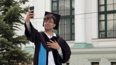 Kepli ve cüppeli bir erkek üniversite binası ve yeşil ağaçların önünde gururla gülümseyen akıllı telefonuyla selfie çeker.