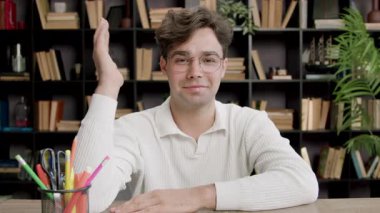 Gözlüklü genç adam, kapalı bir ortamda içi kitaplarla dolu kırtasiye ve kitaplarla dolu bir masada otururken elini kaldırır.