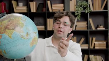 Gözlüklü genç adam kitaplarla dolu bir kütüphane ortamında bir masada otururken çeşitli yerleri gösteren bir küreyle meşgul oluyor.