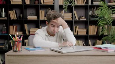 Beyaz kazaklı genç adam masasında hayal kırıklığı yaşıyor, sonra gözleri kapalı, bir çalışma alanında kitaplar ve kırtasiye malzemeleriyle çevrili bir şekilde geriye yaslanıyor.