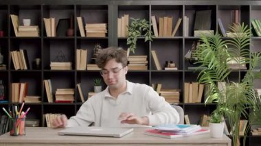 Beyaz kazaklı genç adam dizüstü bilgisayarını kitaplarla ve kırtasiye malzemeleriyle dolu bir masanın üzerine yerleştiriyor.