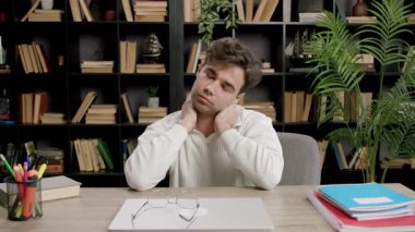 Beyaz kazaklı genç adam boynunu uzatıyor ve sırtına kitaplık takıyor. Gerginlikten ve stresten kurtulmak için görünür bir rahatlama arıyor.