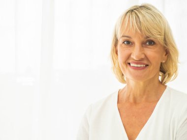 Orta yaşlı güzel bir kadının portresi. Dost canlısı gülümseyen ve oturma odasında kameraya bakan. Kusurlarıyla oynamadan gerçekçi görüntüler..