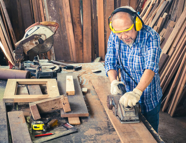 Счастливый старший плотник, использующий электроагрегат с деревянной доской в столярной мастерской. Производство изделий из дерева.