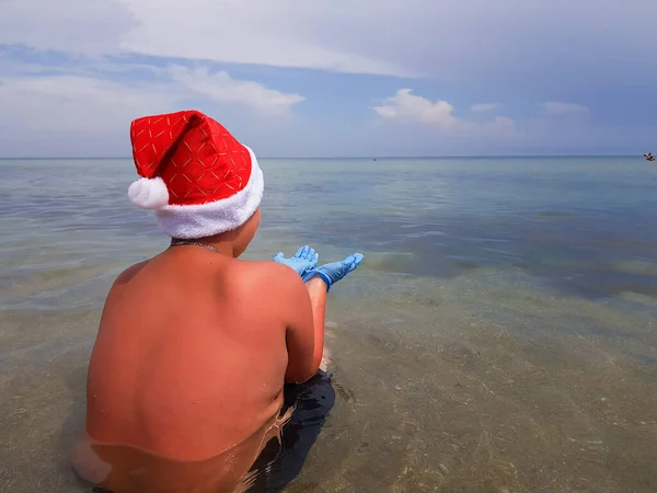 산타클로스가 모자를 의료용 장갑을 해변에 바다를 바라보고 전염병 나기를 기다리고 — 스톡 사진