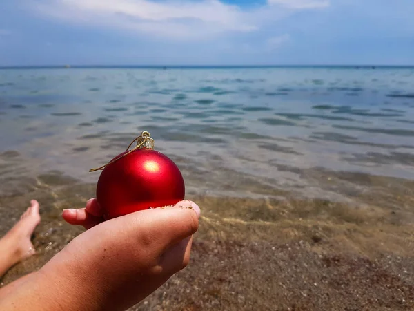 圣诞树玩具的形状是一个红色的球 手里拿着一个小孩背对着大海 圣诞节在海滩上 新年的海洋度假 — 图库照片