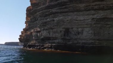 Dağlık bir sahil şeridi. Dik kayalık kayalıkları ve bulutların altında denizin arka planına karşı çalıları var. Video bir tekneden çekilmiş.