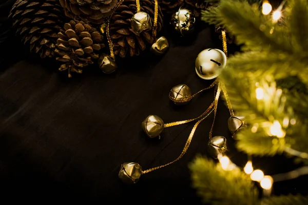 Sinos dourados em um fundo preto com luzes brilhantes de uma grinalda de Natal. Os brinquedos de Natal estão com cones de pinheiro e ramos de abeto verdes. Luzes brilhantes de grinaldas nos ramos de uma árvore de Natal — Fotografia de Stock