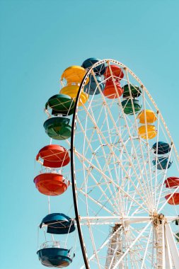 Güneşli bir günde mavi gökyüzü arka planında dönme dolaplar. Gökyüzüne karşı dönme dolabın renkli parlak kabini. Yazın parkta yeşil, sarı, mavi ve kırmızı renkli eğlenceli sürüşler.