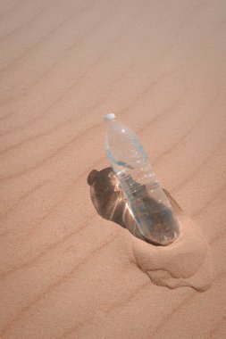 Sıcak bir yaz gününde susuzluğunu gidermek için sarı kumlara yakın bir yerde bir şişe su. Kuma bırakılmış plastik bir şişe soğuk su. Pastel renklerde. Dikey fotoğraf