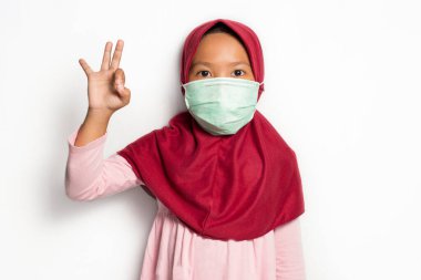 Maske takan küçük Asyalı Müslüman tesettür kızı ve beyaz arka planda baş parmak hareketini göster