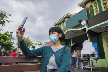Yüz maskeli genç Asyalı kadın halka açık yerlerde cep telefonu selfie 'si kullanıyor. Corona virüsü konsepti.