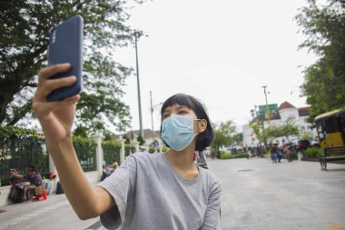 Yüz maskeli genç Asyalı kadın halka açık yerlerde cep telefonu selfie 'si kullanıyor. Corona virüsü konsepti.