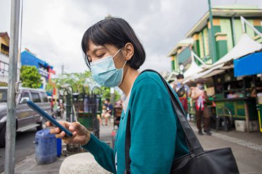 Yüzü maskeli genç Asyalı kadın halka açık yerlerde cep telefonu kullanıyor. Corona virüsü konsepti.