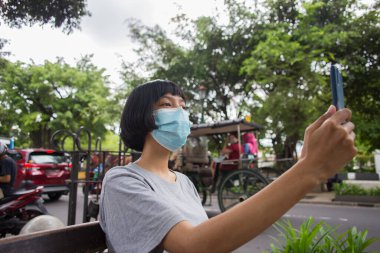 Yüzü maskeli genç Asyalı kadın halka açık yerlerde cep telefonu kullanıyor. Corona virüsü konsepti.