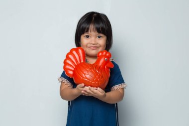 Küçük mutlu Asyalı kız beyaz arka planda izole edilmiş bir tavuk bankasıyla para biriktiriyor.