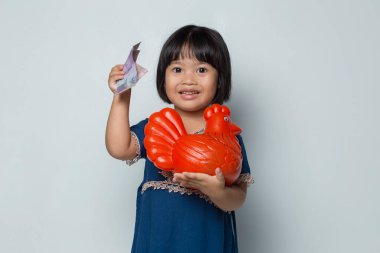 Küçük mutlu Asyalı kız beyaz arka planda izole edilmiş bir tavuk bankasıyla para biriktiriyor.