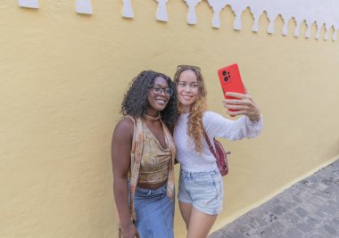 Arkadaşların selfie çekerken çekilmiş yatay bir fotoğrafı. Siyah kız ve sarışın kız..