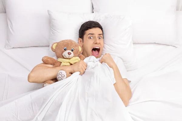 Homem aterrorizado deitado na cama com um urso de pelúcia — Fotografia de Stock