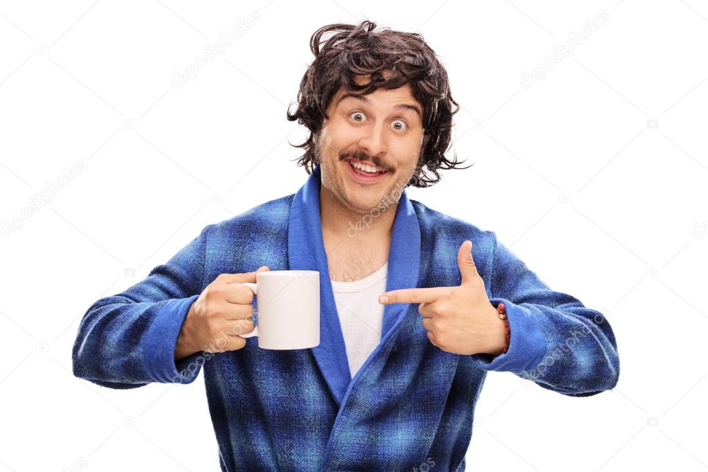 Excited young man in a blue bathrobe holding a ceramic cup and pointing to it with his finger isolated on white background