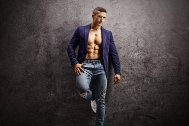 Takım elbiseli yakışıklı bir erkek, üstsüz, paslı gri bir duvara yaslanmış ve kameraya bakıyor.