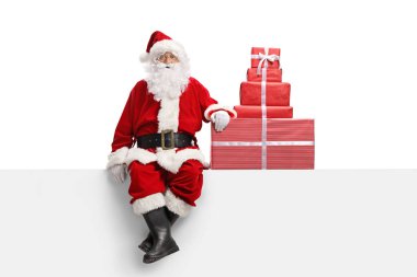Noel Baba beyaz bir pankartta beyaz arka planda izole edilmiş bir yığın Noel hediyesiyle oturuyor.