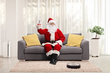 Noel Baba oturma odasındaki bir koltukta oturuyor ve bir robot süpürge halının tozunu alırken yukarı bakıyor. 