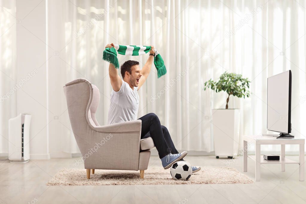 Young man cheering with a scarf and watching a football game on tv at home