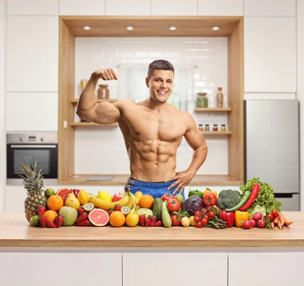 赤身裸体的肌肉男柔韧着二头肌 在厨房柜台上摆出一堆水果和蔬菜的姿势 — 图库照片