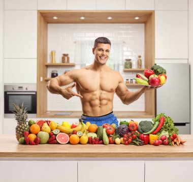 Tabak tutan çıplak erkek manken bir yığın meyve ve sebzeyle mutfakta poz veriyor.